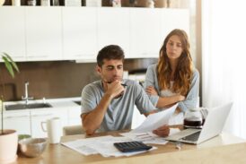 Homme pensif avec lunettes de bureau en main et femme contrariée, assis à une table devant un ordinateur et des documents financiers dans une cuisine lumineuse