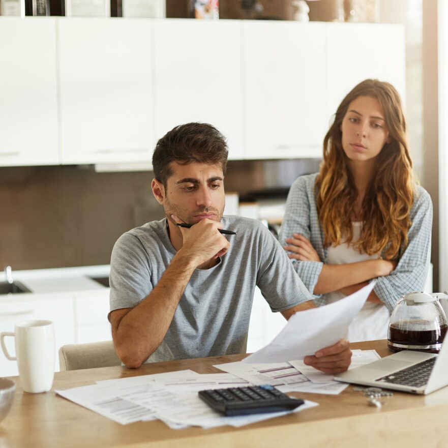 Homme pensif avec lunettes de bureau en main et femme contrariée, assis à une table devant un ordinateur et des documents financiers dans une cuisine lumineuse