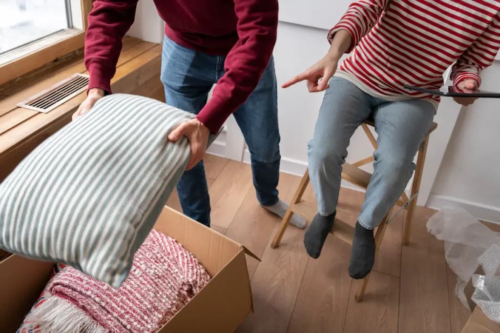 Couple entrain de faire ses cartons de déménagement. L'homme est debout avec un coussin en main qu'il met dans un carton. La femme est assise sur un tabouret avec une tablette en main et semble lui indiquer dans quel carton mettre les affaires.