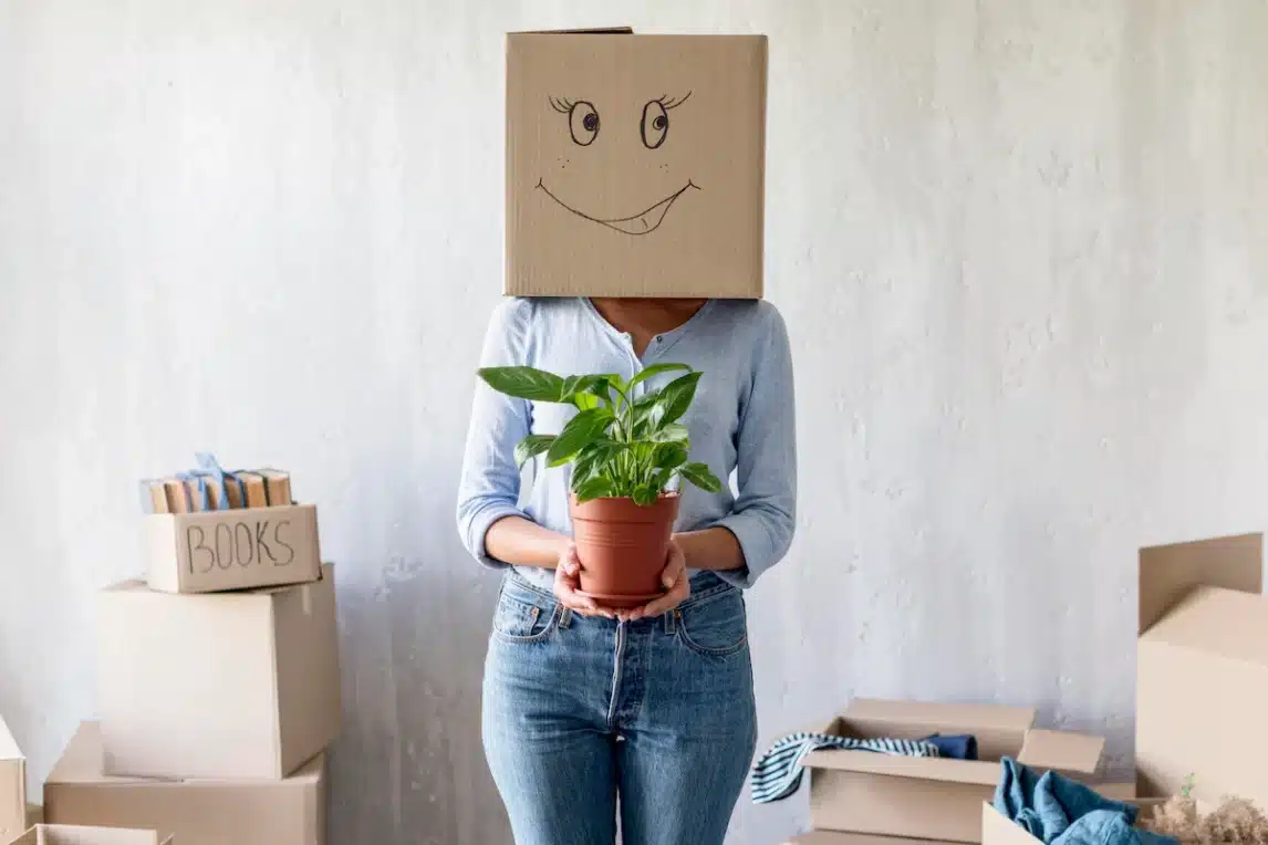 Dame avec un carton sur la tête sur lequel est représenté un emoji qui sourit. La dame tient dans ses mains un pot avec une plante verte, pour faire le liens avec un déménagement écologique.