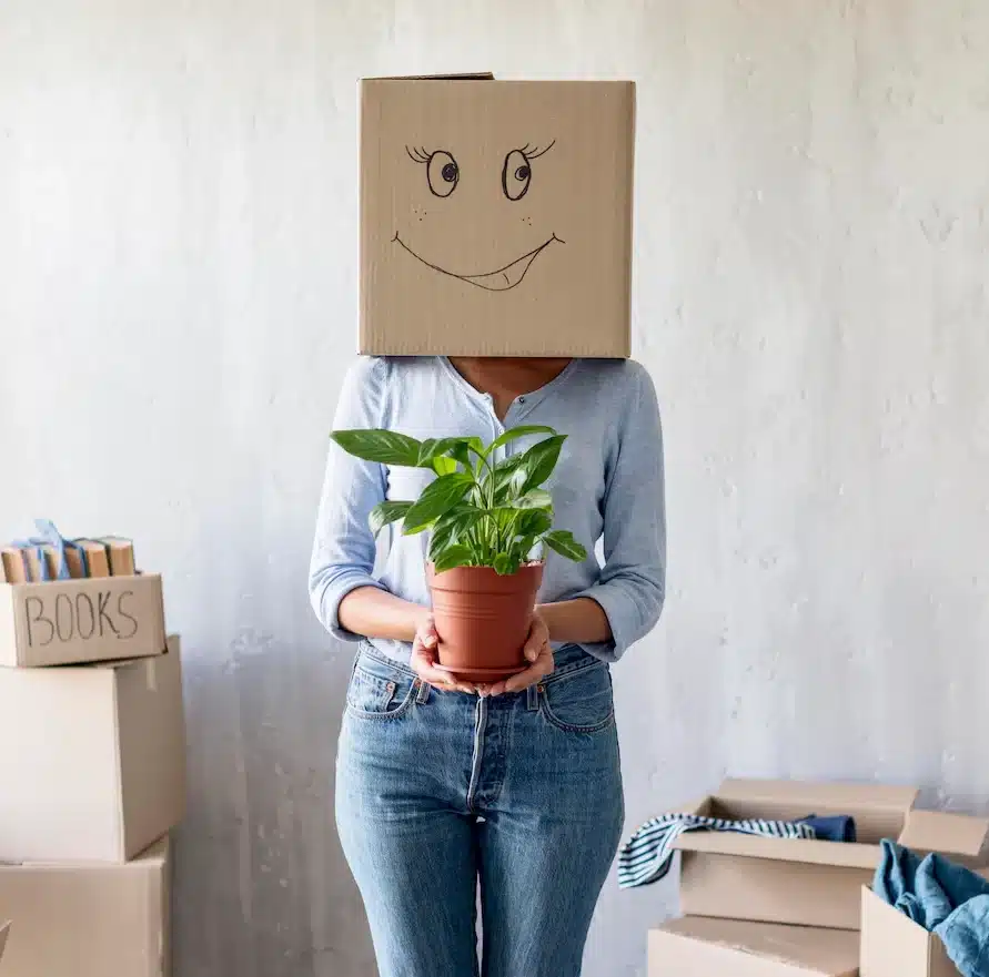 Dame avec un carton sur la tête sur lequel est représenté un emoji qui sourit. La dame tient dans ses mains un pot avec une plante verte, pour faire le liens avec un déménagement écologique.