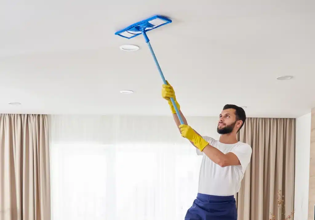 Homme debout avec un balais entrain de nettoyer le plafond d'un appartement.