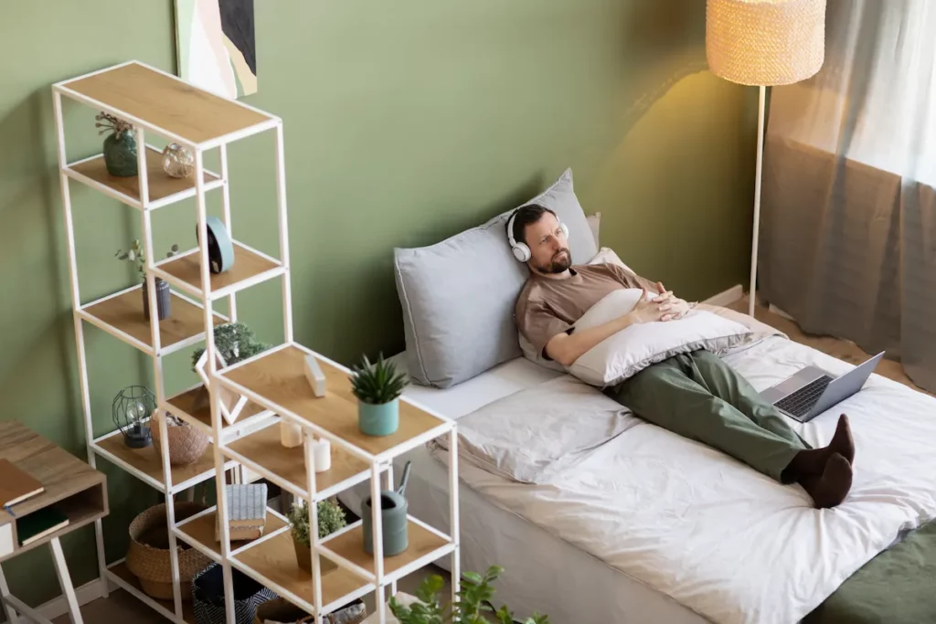 Un homme allongé sur sont lit dans une chambre. Il semble être entrain d'écouter de la musique avec son casque sur les oreilles et son ordinateur à proximité. 