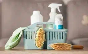 Panier bleu rempli de produits ménagers et d’accessoires de nettoyage, avec une brosse, une éponge et des flacons de savon, posés sur une table.