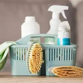 Panier bleu rempli de produits ménagers et d’accessoires de nettoyage, avec une brosse, une éponge et des flacons de savon, posés sur une table.