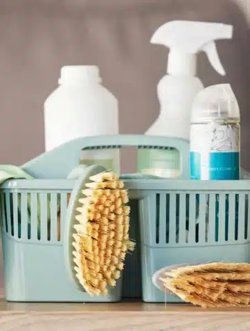Panier bleu rempli de produits ménagers et d’accessoires de nettoyage, avec une brosse, une éponge et des flacons de savon, posés sur une table.