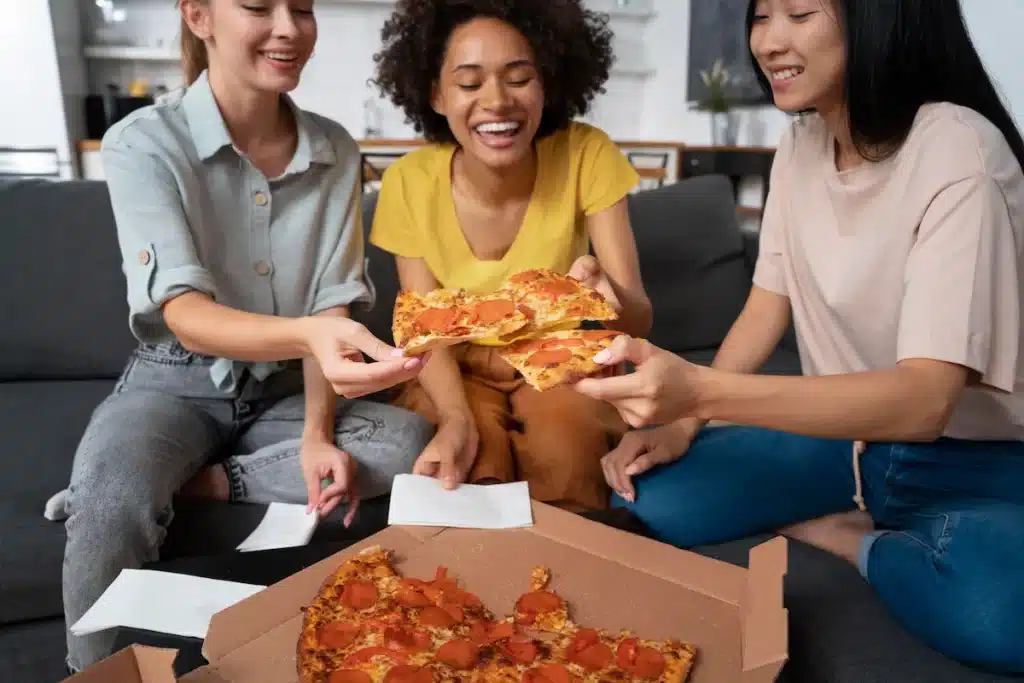 3 colocataires, entrain de manger une pizza. Elles sont toutes les 3 assises sur un canapé, avec un part de pizza en main.