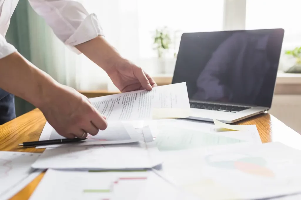 Documents administratifs étalés sur un bureau avec un ordinateur et les mains d'un homme qui tient une feuille de papier.