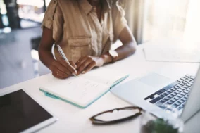 Personne assise à un bureau, écrivant sur un carnet ouvert avec un stylo, ordinateur portable et lunettes posés à côté.