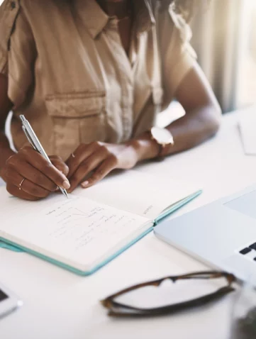Personne assise à un bureau, écrivant sur un carnet ouvert avec un stylo, ordinateur portable et lunettes posés à côté.