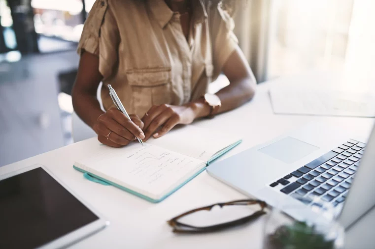 Personne assise à un bureau, écrivant sur un carnet ouvert avec un stylo, ordinateur portable et lunettes posés à côté.