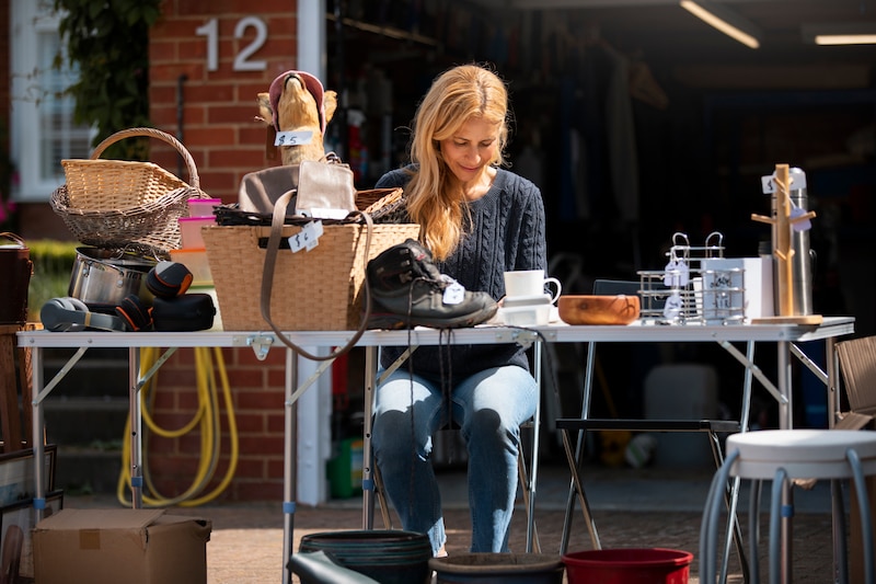 Femme assise derrière sa table de brocante, vendant des chaussures, paniers en osier et objets de cuisine.