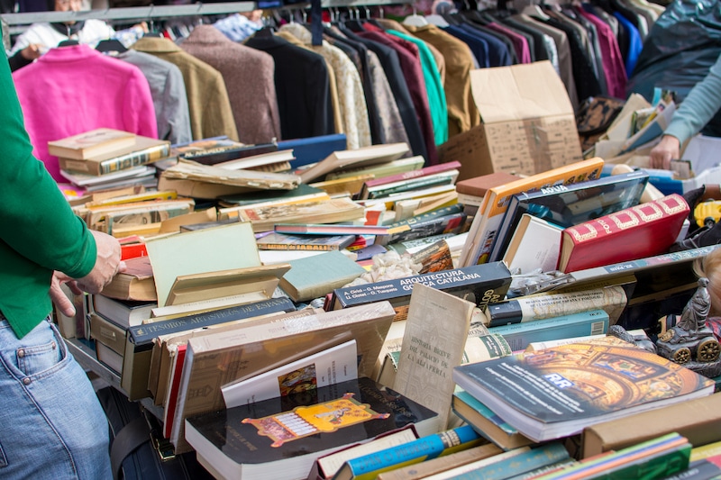 Femme cherchant des livres d'occasion dans une brocante, avec des livres en désordre au premier plan et des vestes colorées suspendues sur un portant au second plan.