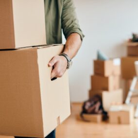 Un déménageur tire un grand carton de l’intérieur d’un logement, avec d’autres cartons et du matériel de bricolage flous en arrière-plan.