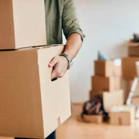 Un déménageur tire un grand carton de l’intérieur d’un logement, avec d’autres cartons et du matériel de bricolage flous en arrière-plan.