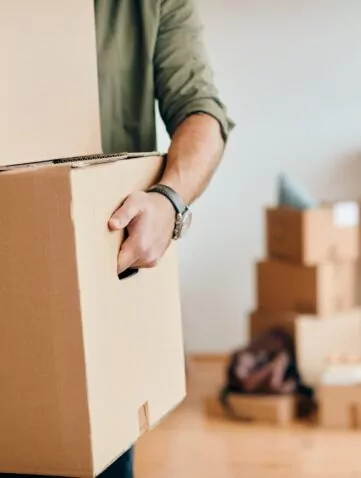 Un déménageur tire un grand carton de l’intérieur d’un logement, avec d’autres cartons et du matériel de bricolage flous en arrière-plan.