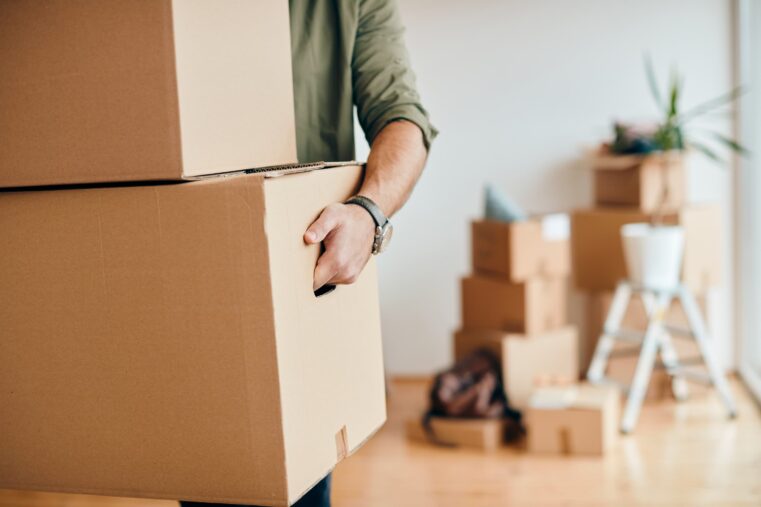 Un déménageur tire un grand carton de l’intérieur d’un logement, avec d’autres cartons et du matériel de bricolage flous en arrière-plan.