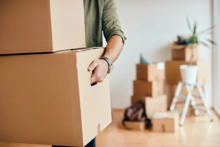 Un déménageur tire un grand carton de l’intérieur d’un logement, avec d’autres cartons et du matériel de bricolage flous en arrière-plan.