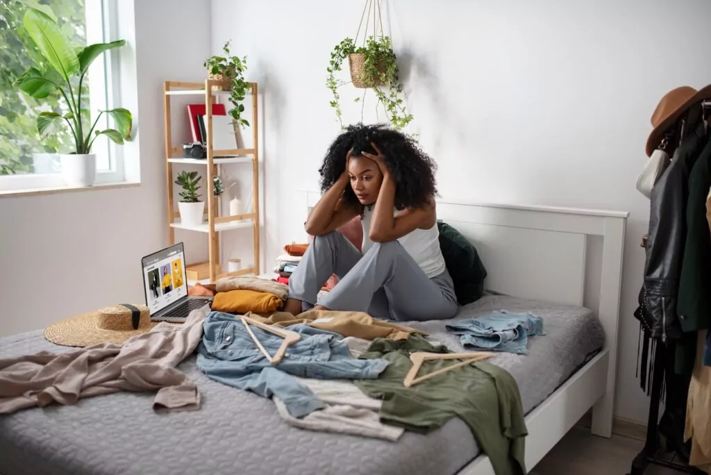 Femme qui est assise sur son lit désespérée que sa chambre soit en désordre et veut désencombrer son armoire 